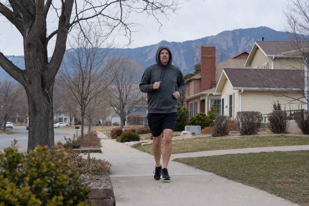 A single casual runner in a hoodie jogging at an easy pace along a suburban sidewalk on a calm morning, foothills in the distance