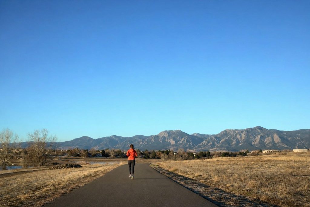 A lone runner seen from behind on an open paved road heading toward the Front Range under a vast pale-blue Colorado sky
