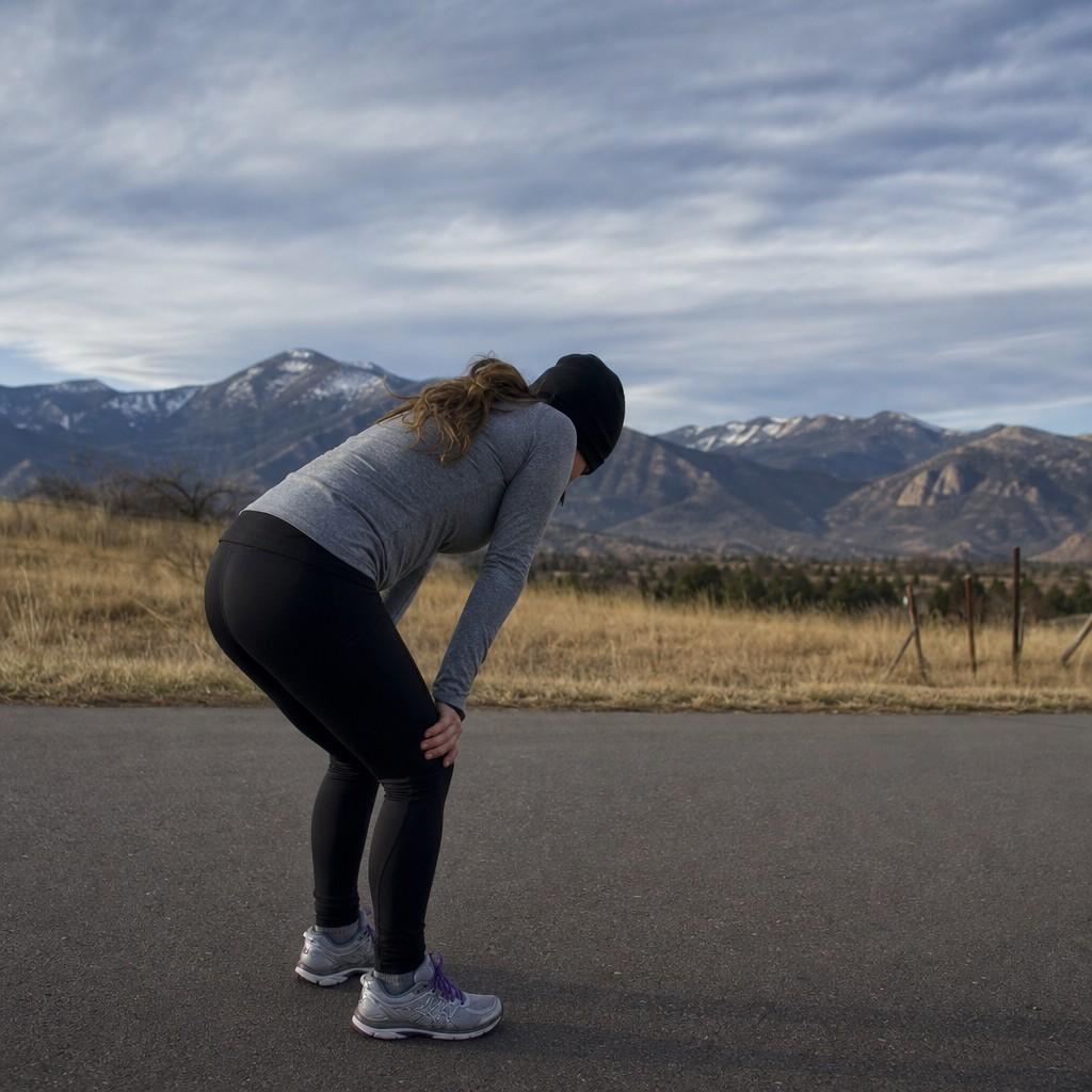 A runner bent over with hands on knees catching their breath on an open path with mountains behind
