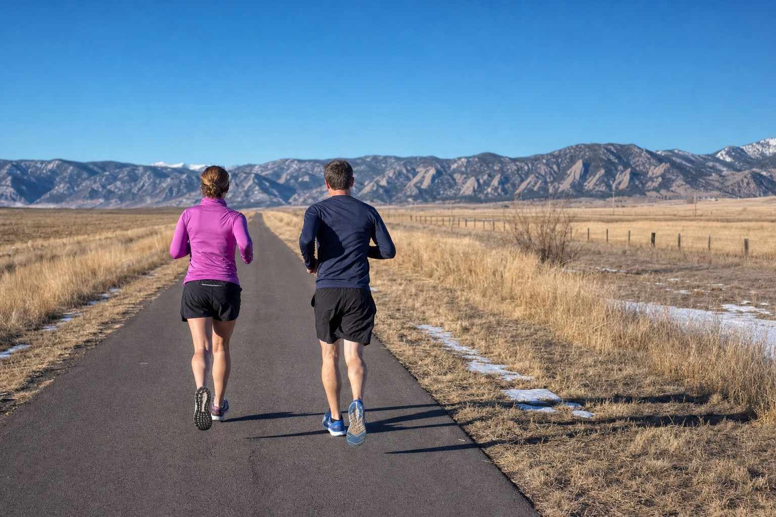 Two runners seen from behind on a long open paved path stretching toward the Front Range, a steady long-run feel