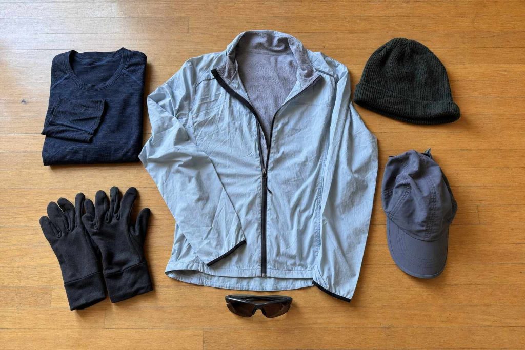 An overhead flat-lay of layered running clothes for Colorado: a base layer, a light shell, gloves, a knit hat, a cap and sunglasses laid out on a wood floor