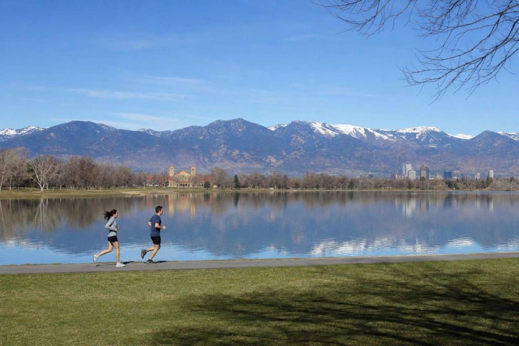 A wide view of two small runners on a flat paved path beside a city lake on a clear morning, the Front Range reflected in the water