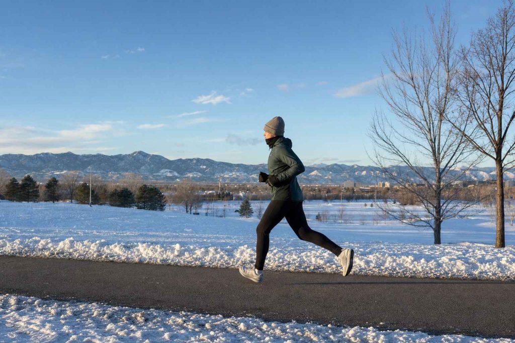 A single runner in winter layers running along a snow-edged park road on a clear cold Denver morning, breath visible, foothills behind