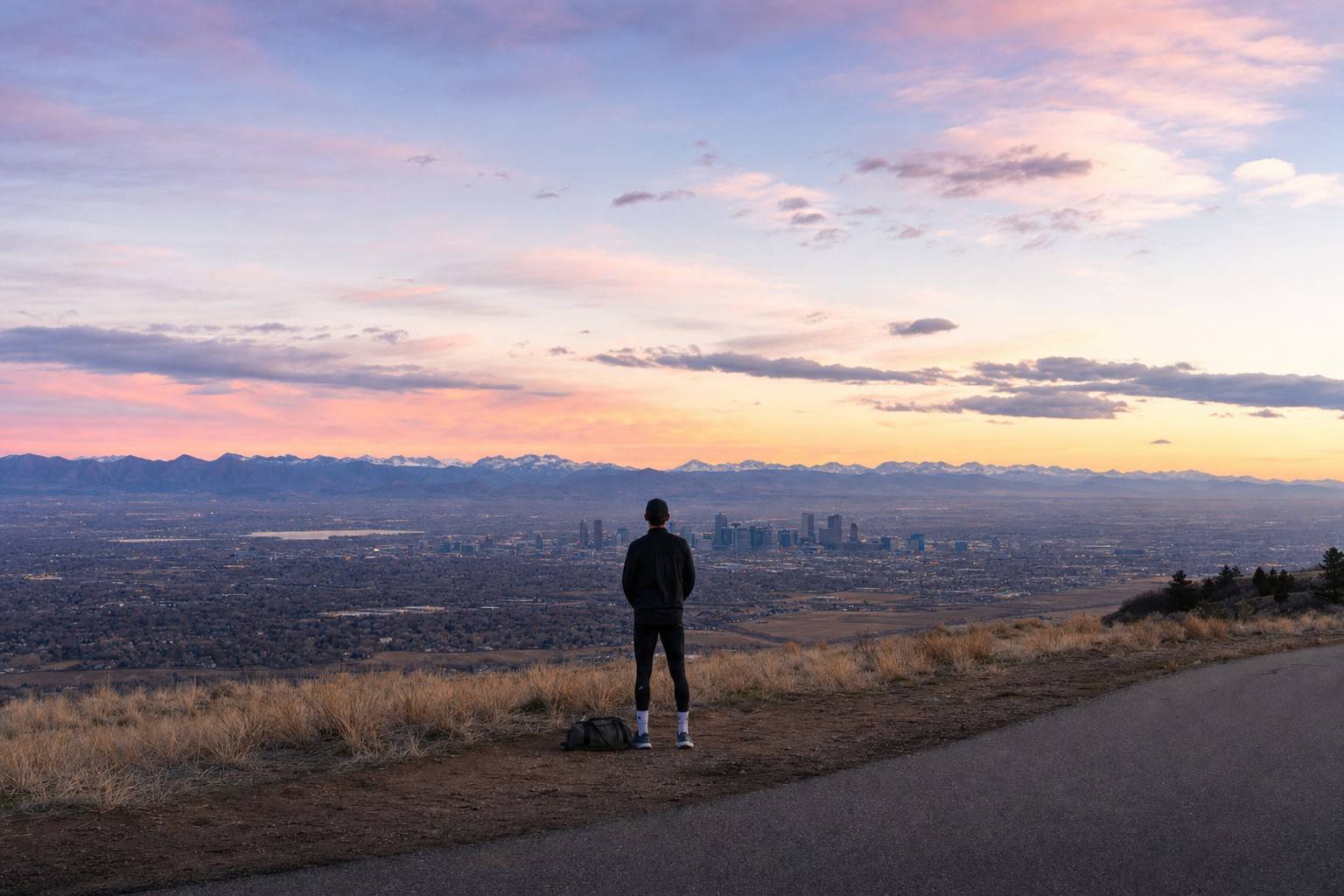 A lone runner with a small travel duffel standing at a high overlook at dawn, the Denver skyline distant below and mountains on the horizon