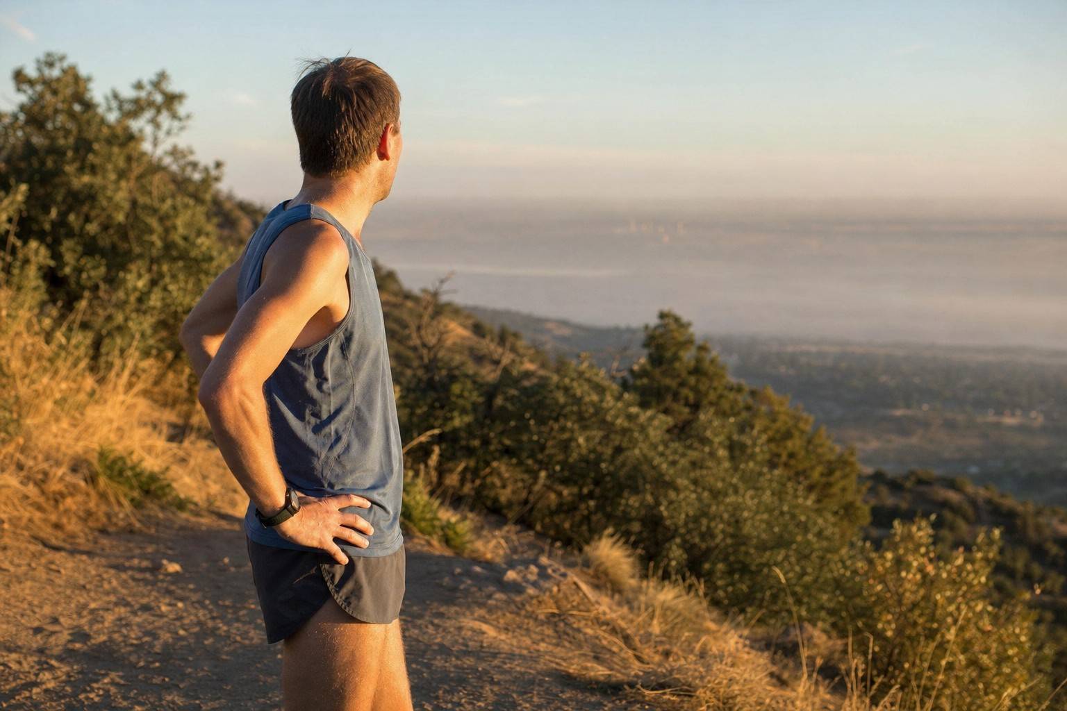 A runner in a tank top pausing on a dry foothills trail at sunrise, with a hazy layer sitting over the plains below