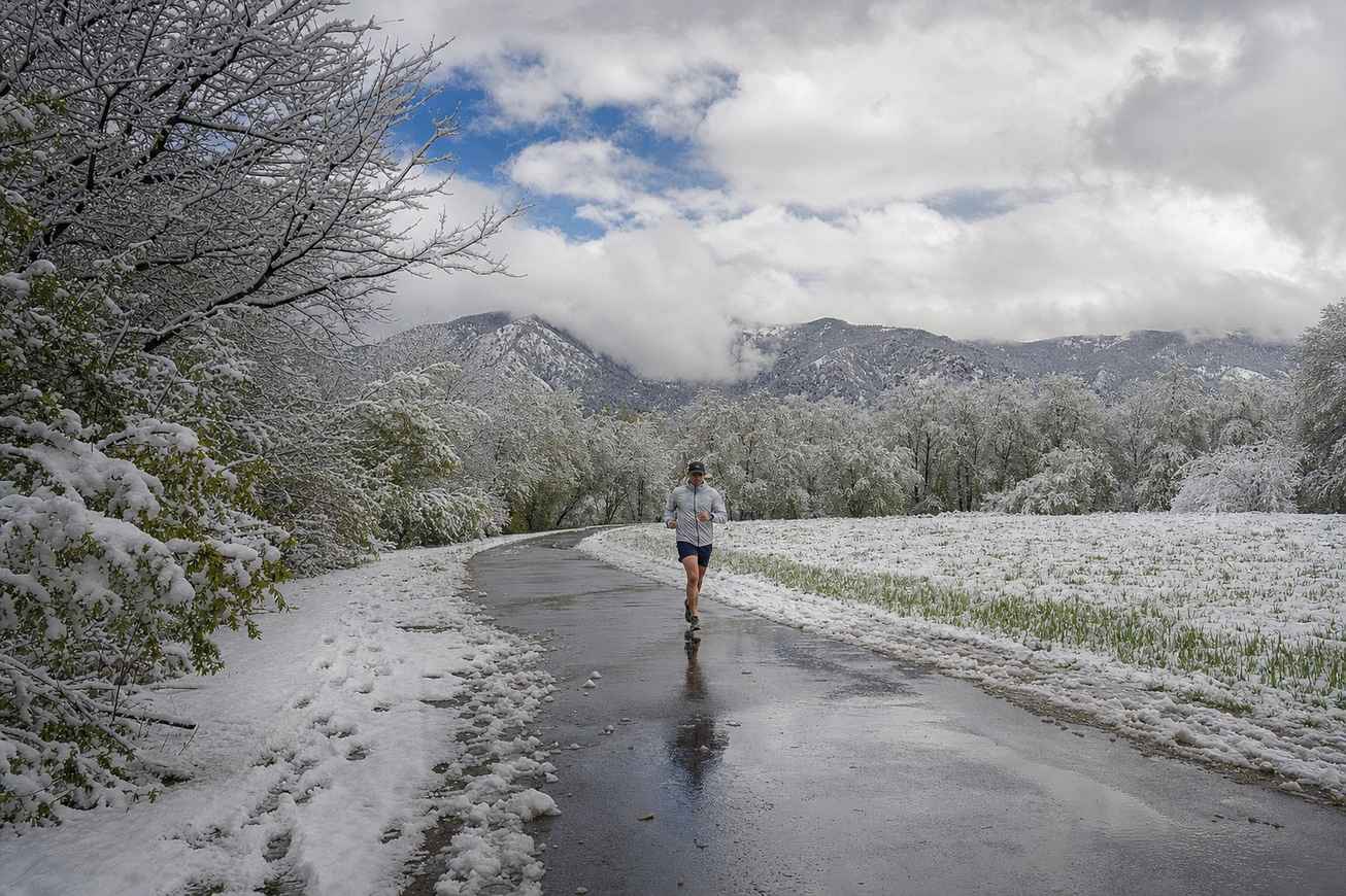 A runner in a light shell jacket coming toward the camera along a slushy park path under heavy wet spring snow