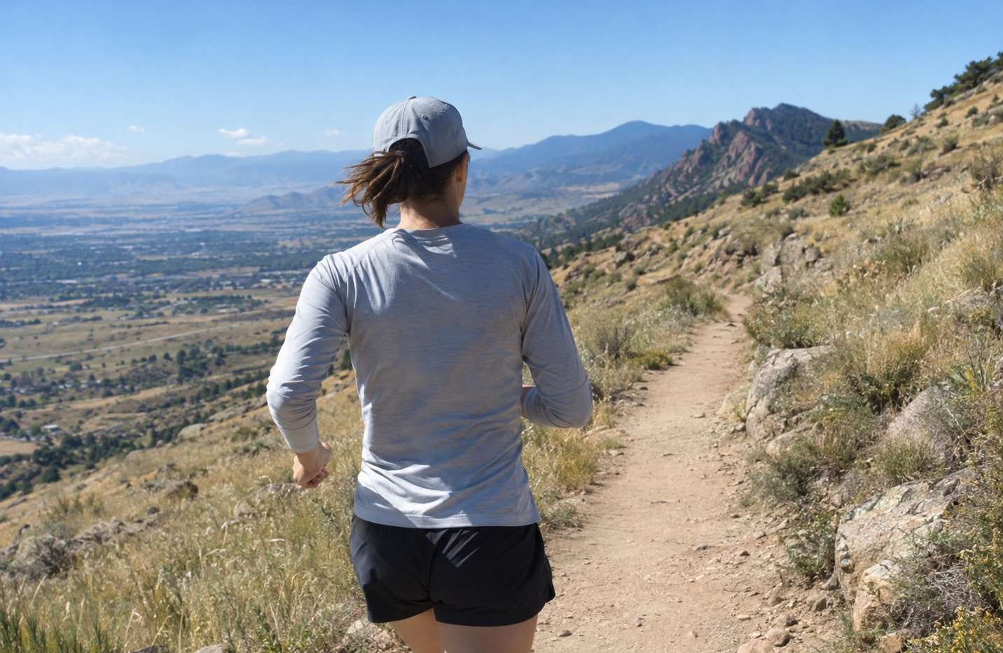 A runner seen from behind on an open ridgeline trail under a hard high-altitude sun, shoulders and neck covered by a light long sleeve and a brimmed cap
