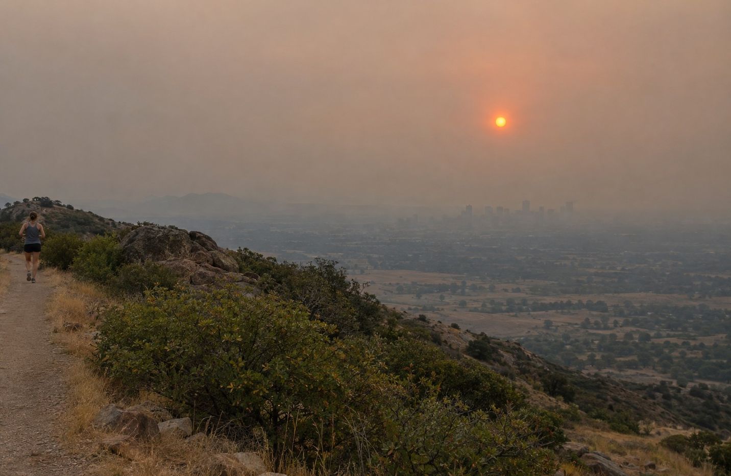The Denver skyline flattened into grey haze under wildfire smoke, seen from a foothills overlook with the sun a dull orange disc